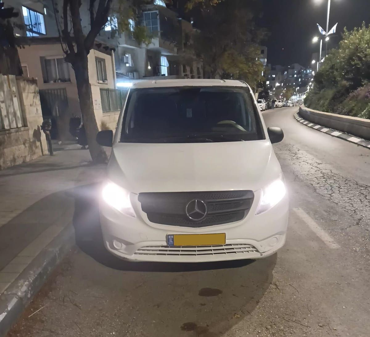 חזית של ואן מרצדס לבן עם פנסים דולקים בלילה, ממתין לנוסעים עבור הסעות לנתבג. (מתאר ואן לבן חונה בלילה)