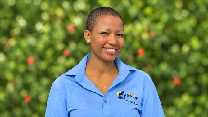 Smiling person in a tan cap and blue shirt standing outdoors with mountains in the background