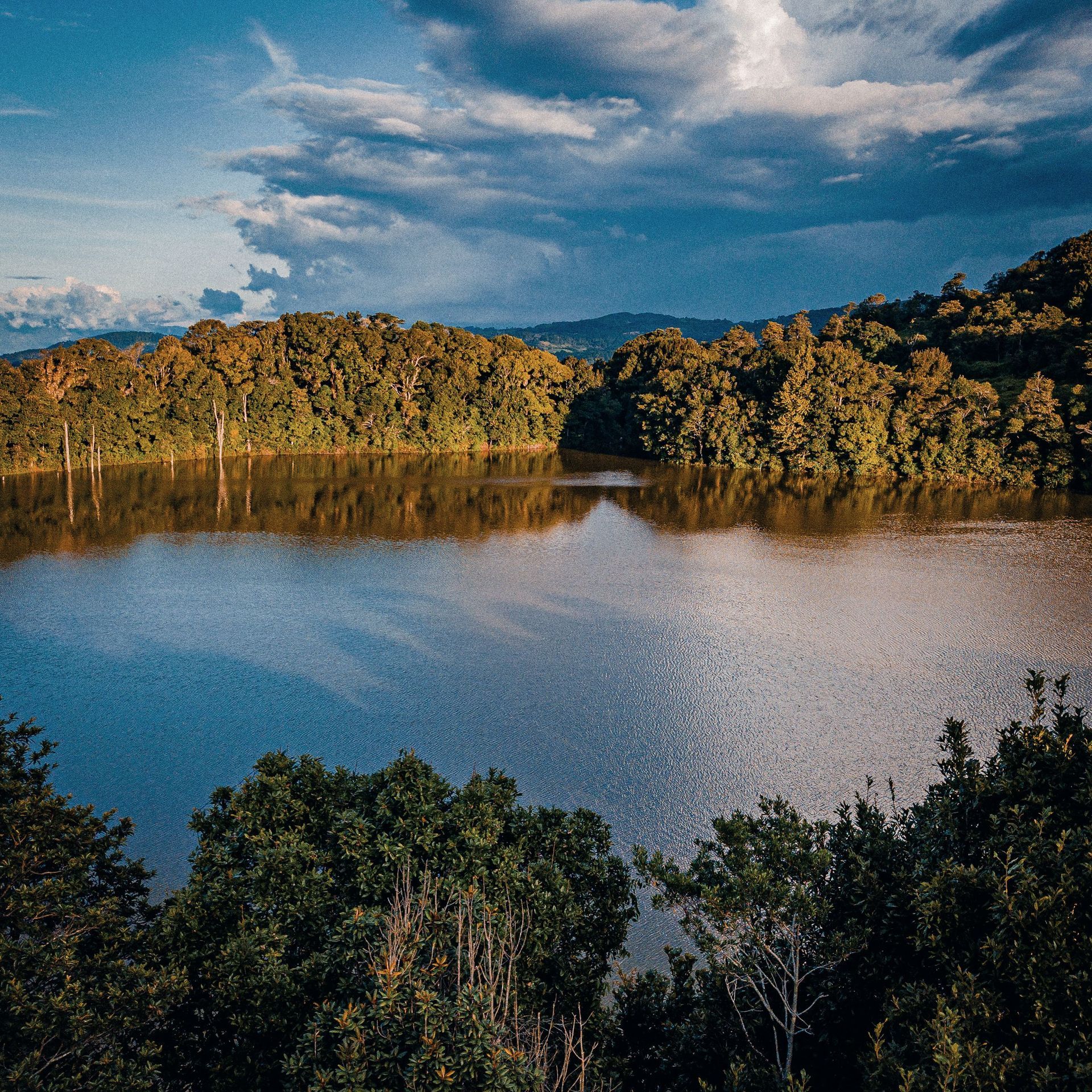 A calm lake reflecting golden forested hills under a blue, partly cloudy sky