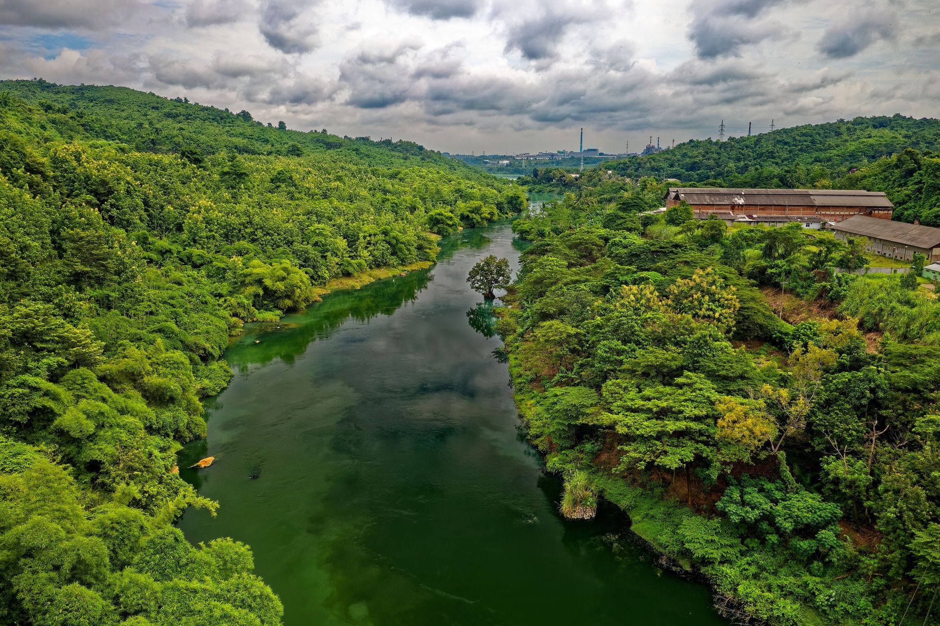 Lush green river valley beneath cloudy sky, with buildings along the right bank