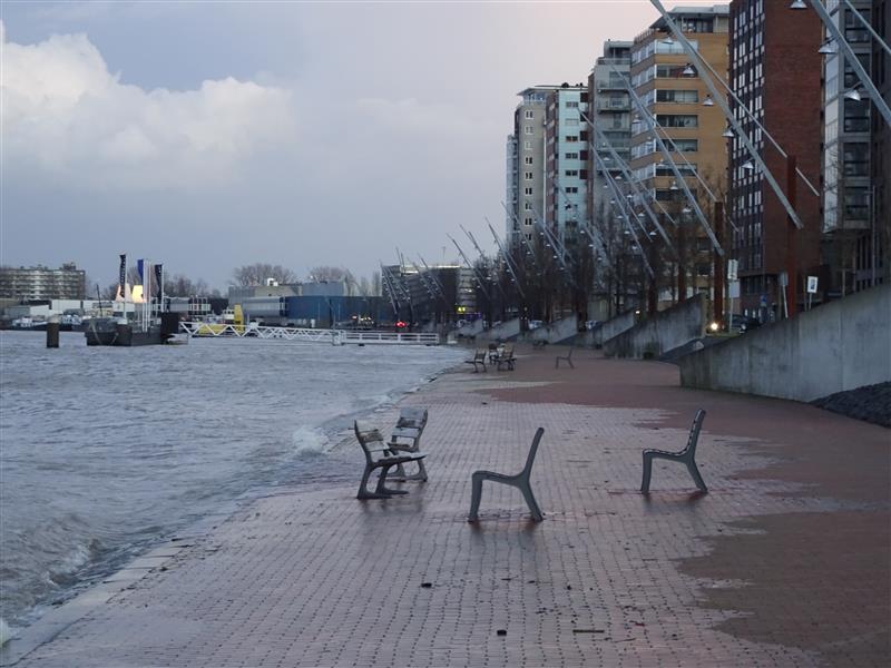 Drie moderne stoelen op een bakstenen oever, golven die vlakbij tegen de kust slaan, stadsgebouwen op de achtergrond onder een bewolkte hemel.