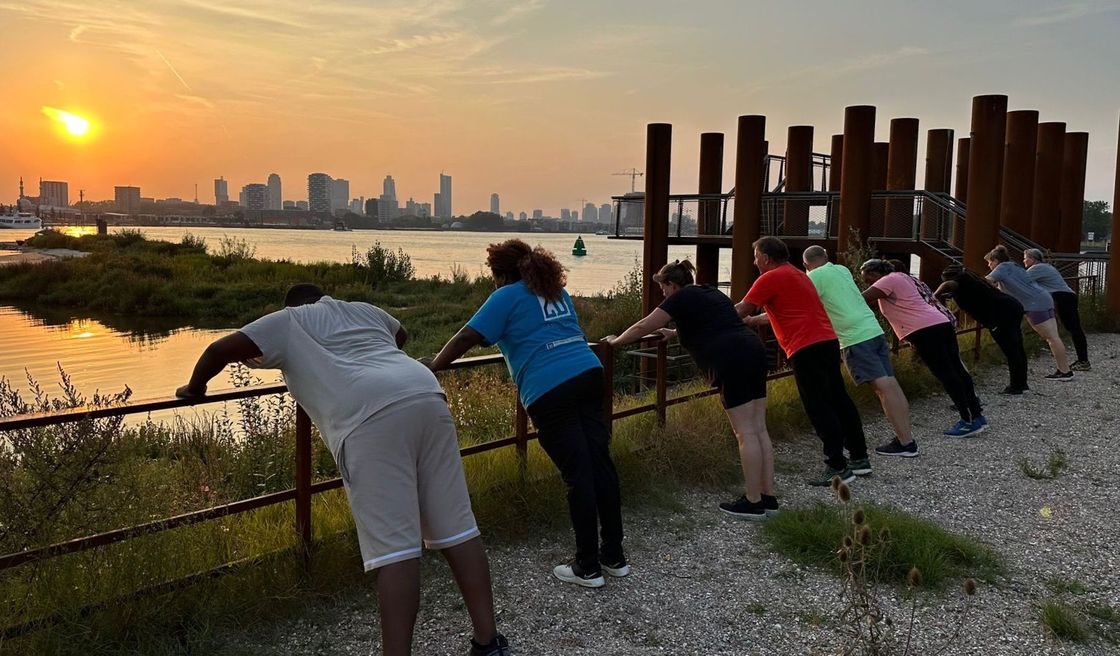 Mensen doen push-ups op een helling bij zonsondergang, met uitzicht op een wateroppervlak en de skyline van de stad.
