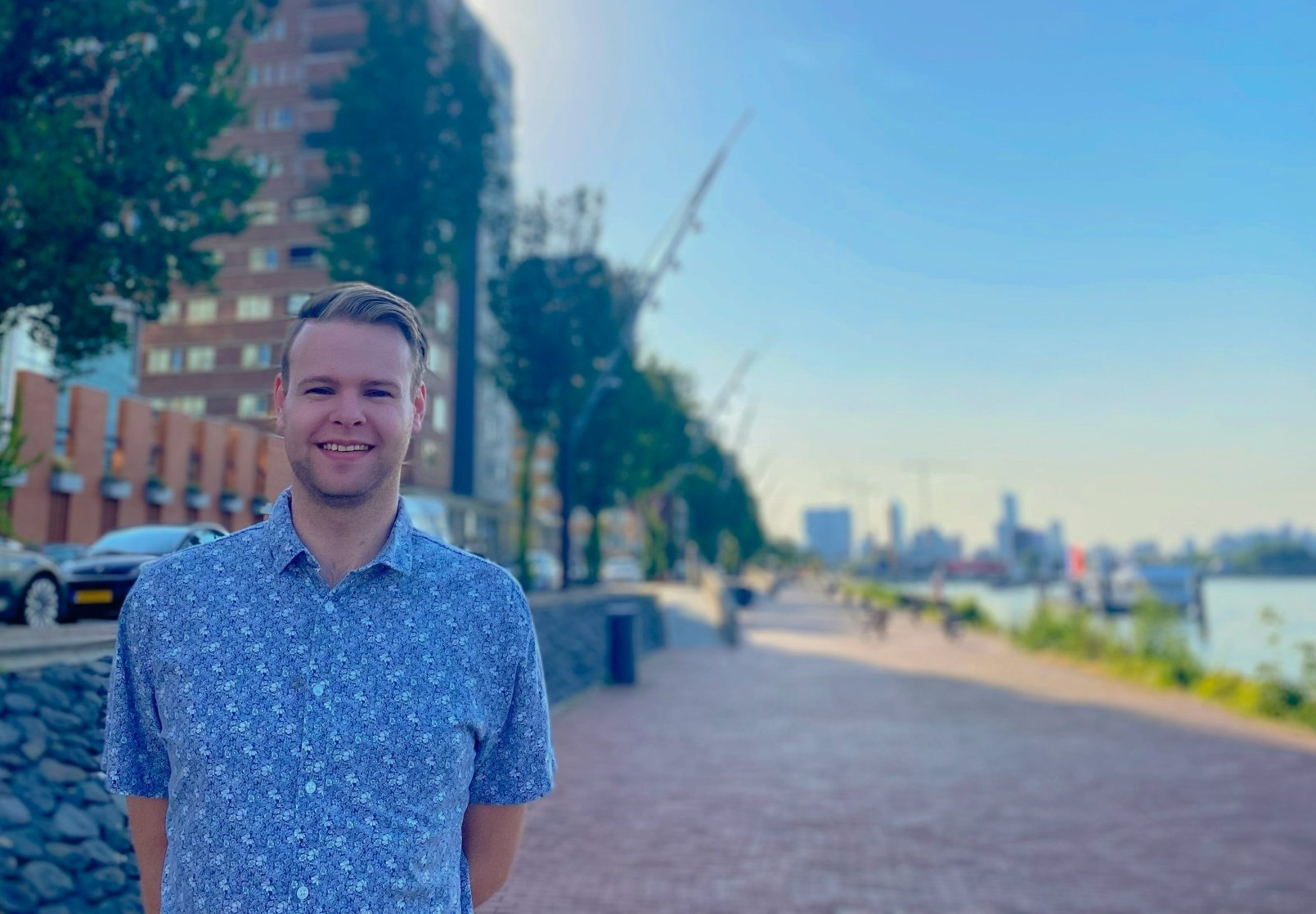Een man in een blauw overhemd met patroon lacht buiten, vlakbij een wandelpad langs het water met uitzicht op de skyline van de stad.