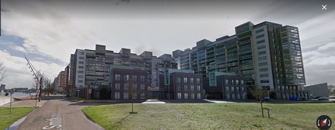 Gebouwen langs een wandelpad aan het water, met een grasveld op de voorgrond en een bewolkte hemel.
