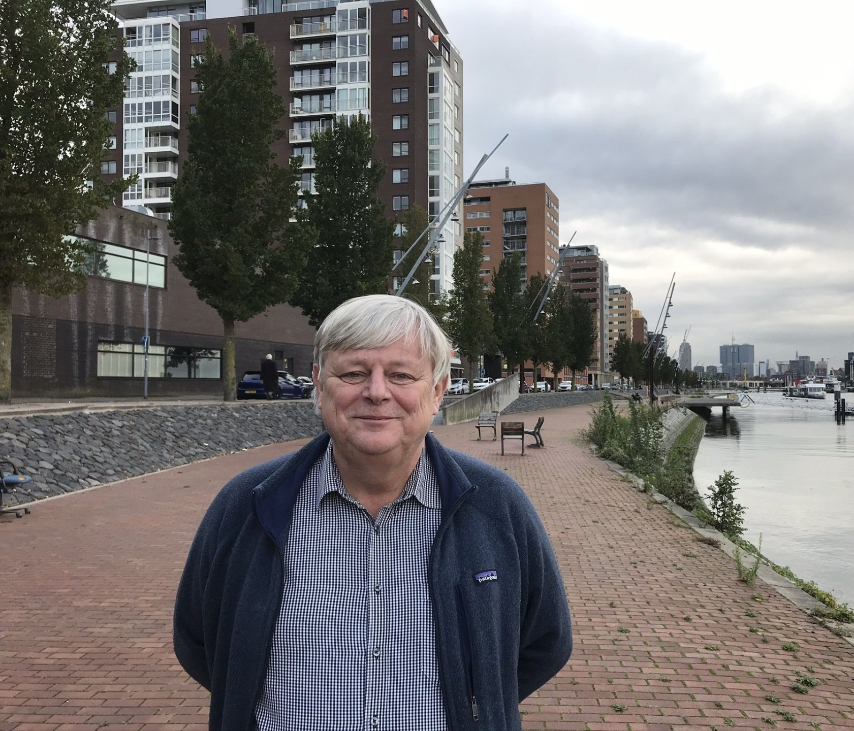 Een man in een blauwe jas staat op een stenen pad langs een kanaal met gebouwen op de achtergrond, op een bewolkte dag.