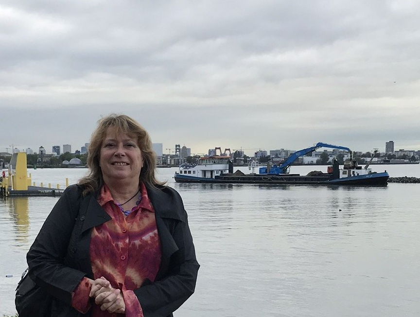 Vrouw voor het water en een binnenschip met een stadsgezicht op de achtergrond; bewolkte dag.