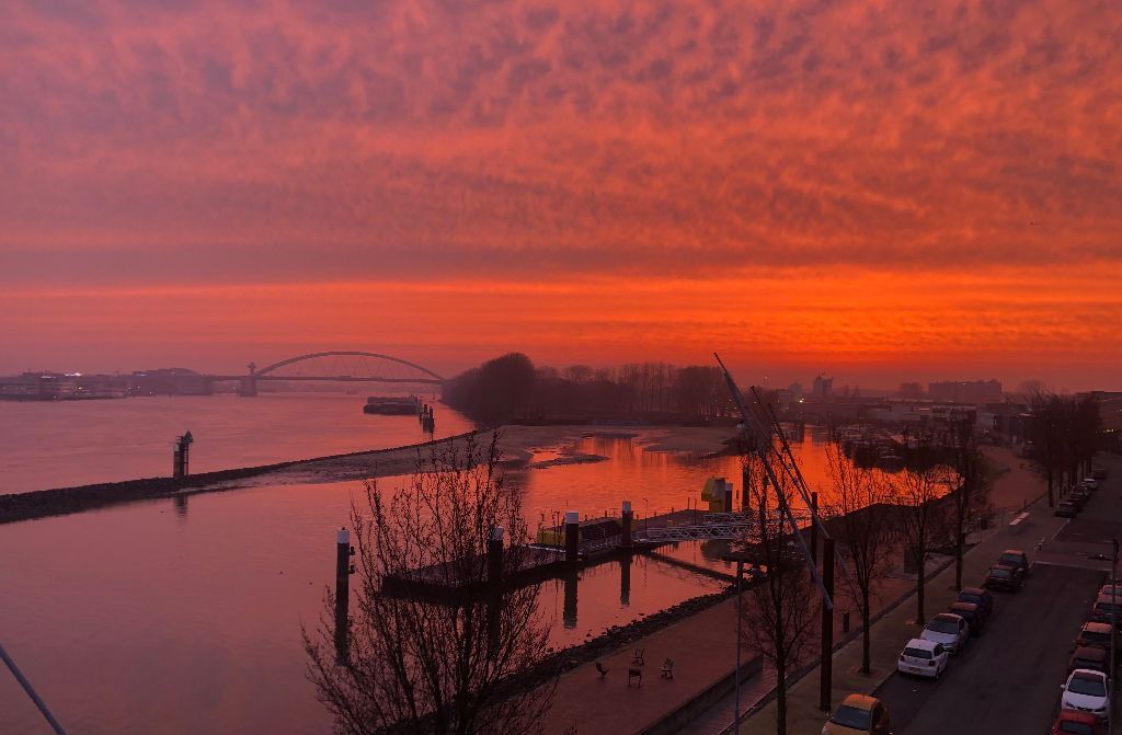 Zonsondergang boven de rivier, met een oranje en roze lucht die in het water weerspiegelt; een brug in de verte, steigers en bomen op de voorgrond.