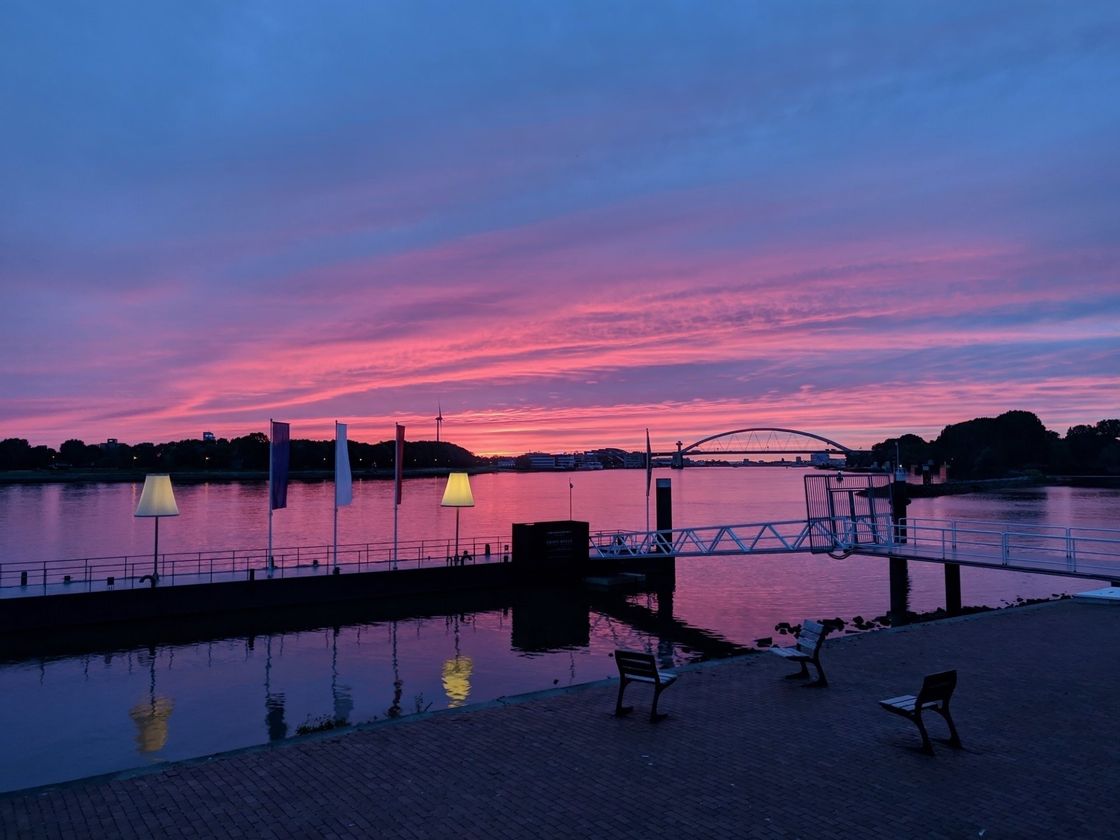 Zonsondergang boven een rivier met roze en paarse tinten. Een steiger met bankjes en verlichting; een brug in de verte.