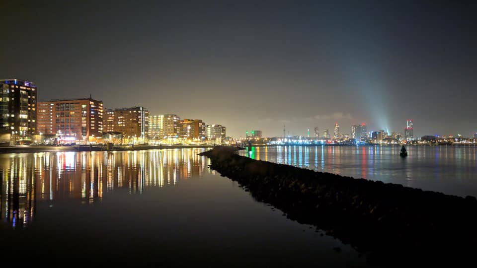 Een nachtelijk stadsgezicht dat weerspiegeld wordt in het kalme water, met een lange golfbreker en verlichte gebouwen.