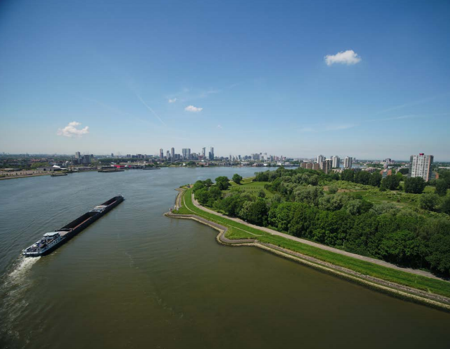 Een vrachtschip vaart over een brede rivier met in de verte een stadsgezicht onder een blauwe hemel.