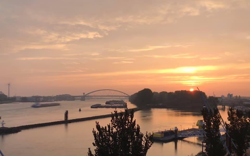 Zonsondergang boven een rivier met een brug, boten en silhouetten van bomen. Oranje en gouden tinten domineren de lucht en het water.