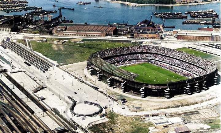 Luchtfoto van een volgepakt stadion naast een haven en spoorlijnen. Groen veld, bruine gebouwen en blauw water.