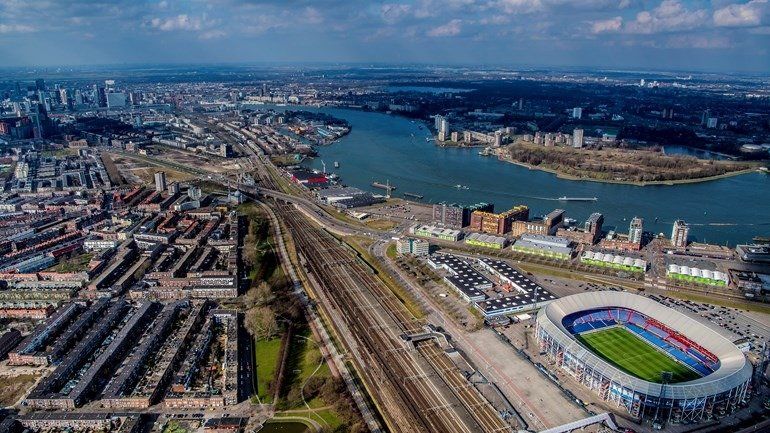 Luchtfoto van een stad met een rivier. Op de voorgrond is een stadion te zien, met spoorlijnen en gebouwen.