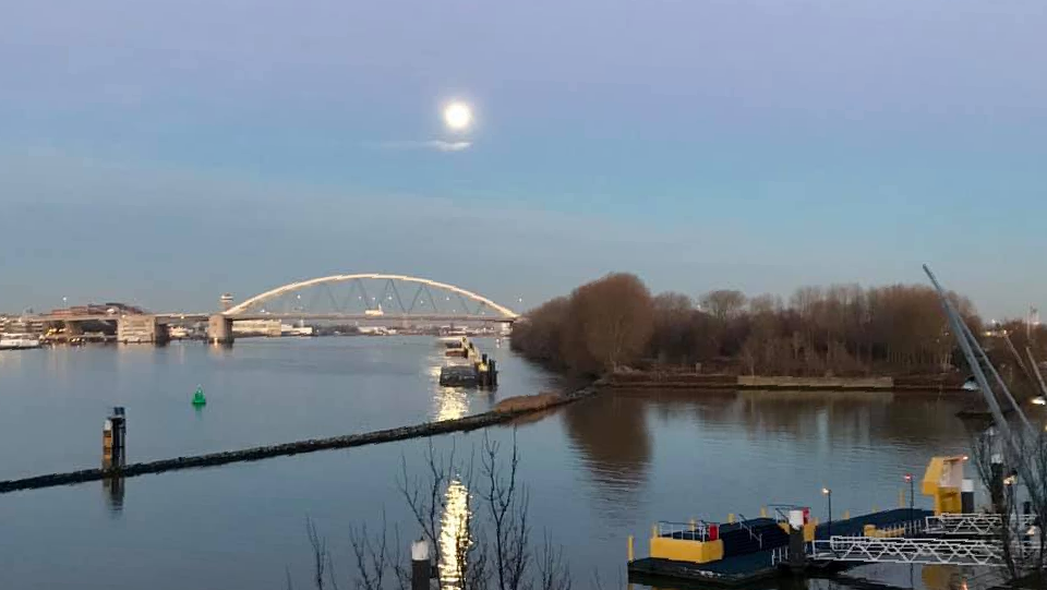Een rivier met een brug, een boot en de maan aan de schemering.