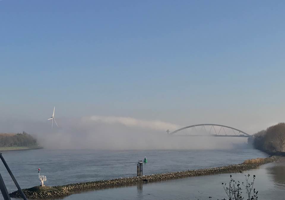 Een rivier met een brug en een windmolen, gedeeltelijk aan het zicht onttrokken door mist, onder een blauwe hemel.