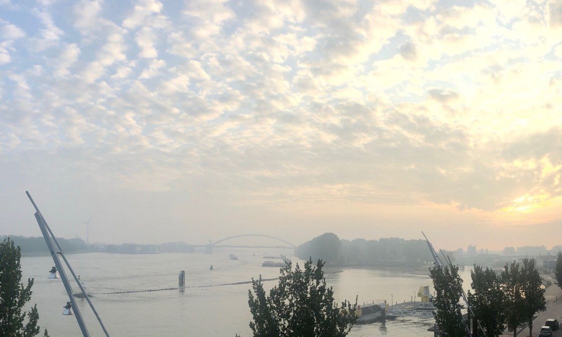 Rivierlandschap met bewolkte hemel bij zonsopgang. Bomen en een brug in de verte, zacht licht.