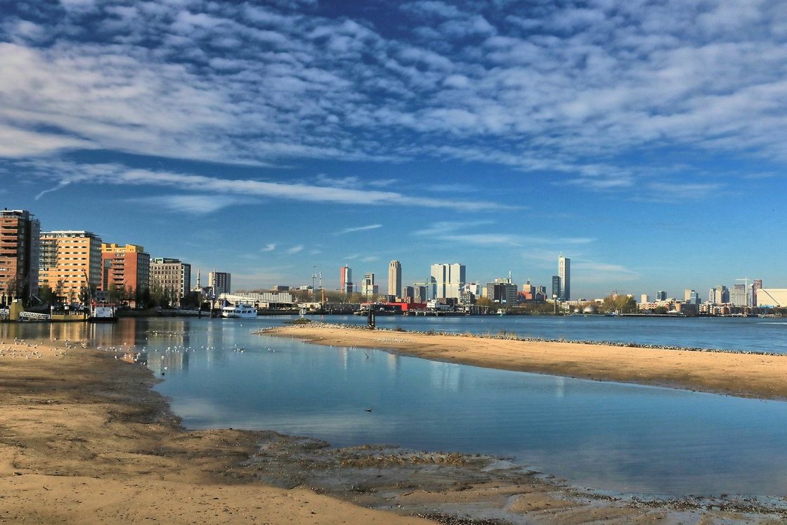 De skyline van de stad aan de overkant van kalm water, met het strand op de voorgrond onder een gedeeltelijk bewolkte blauwe hemel.