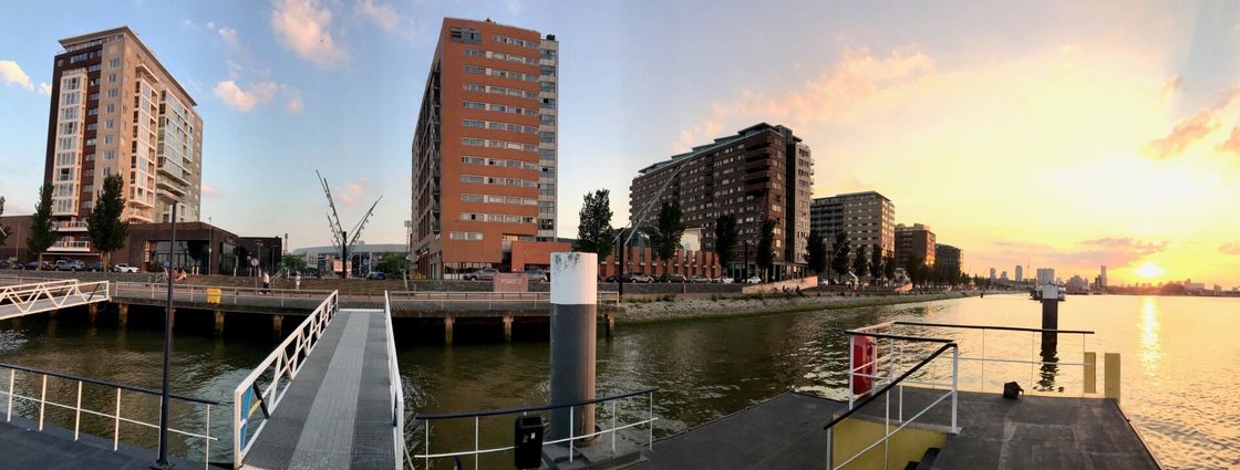 Panoramisch uitzicht op gebouwen aan het water tijdens zonsondergang. Een pier leidt naar het water met een kleurrijke hemel op de achtergrond.