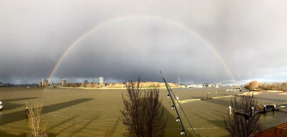 Een complete regenboog boven een wateroppervlak en een stadsgezicht in de verte; een grijze, bewolkte lucht.