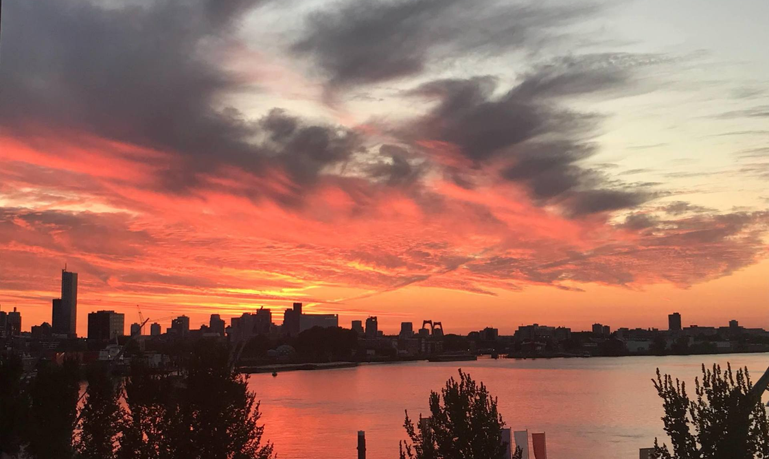 Zonsondergang boven de skyline van de stad, weerspiegeld in het water, met oranje en grijze wolken.