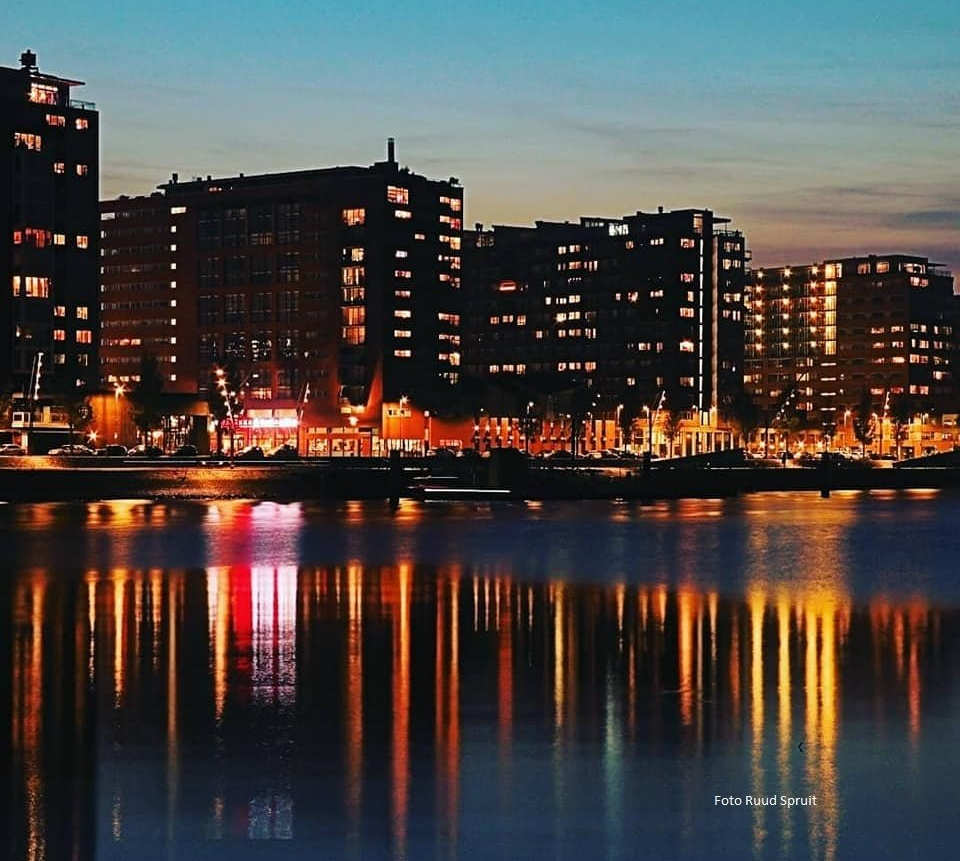 De skyline van de stad weerspiegelt zich in het water bij schemering. Gebouwen met verlichte ramen, oranje en blauwe tinten.