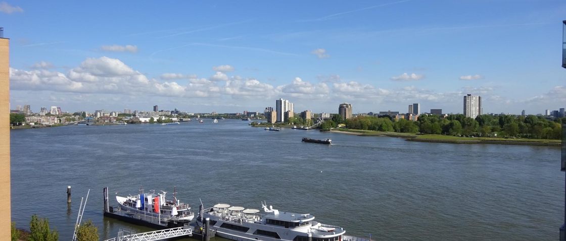 Uitzicht vanaf het water op de skyline van een stad onder een blauwe hemel met wolken. Op de voorgrond ligt een boot aangemeerd.