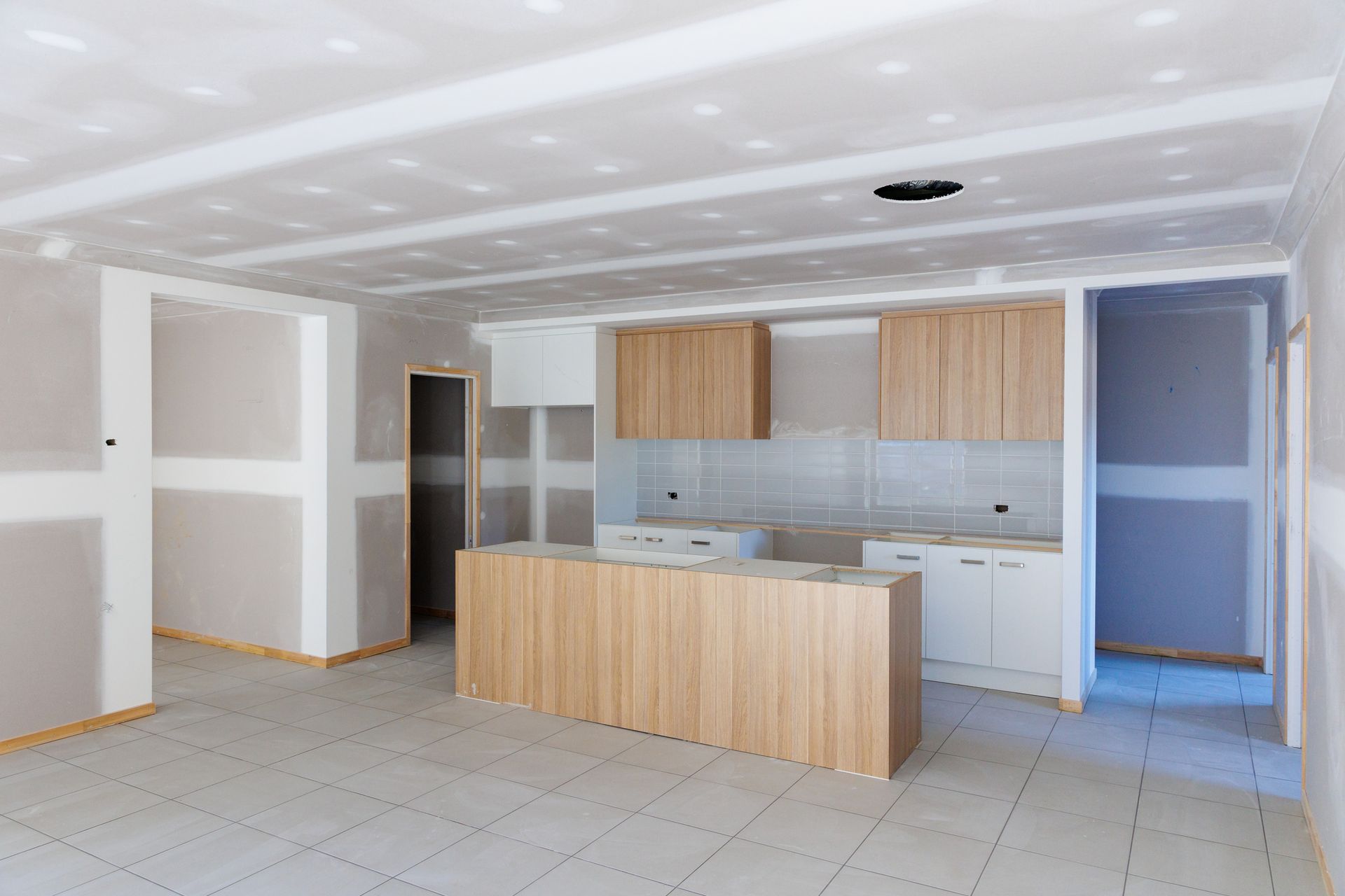 Kitchen under construction with wood cabinets and island, drywall walls, and tile floor.