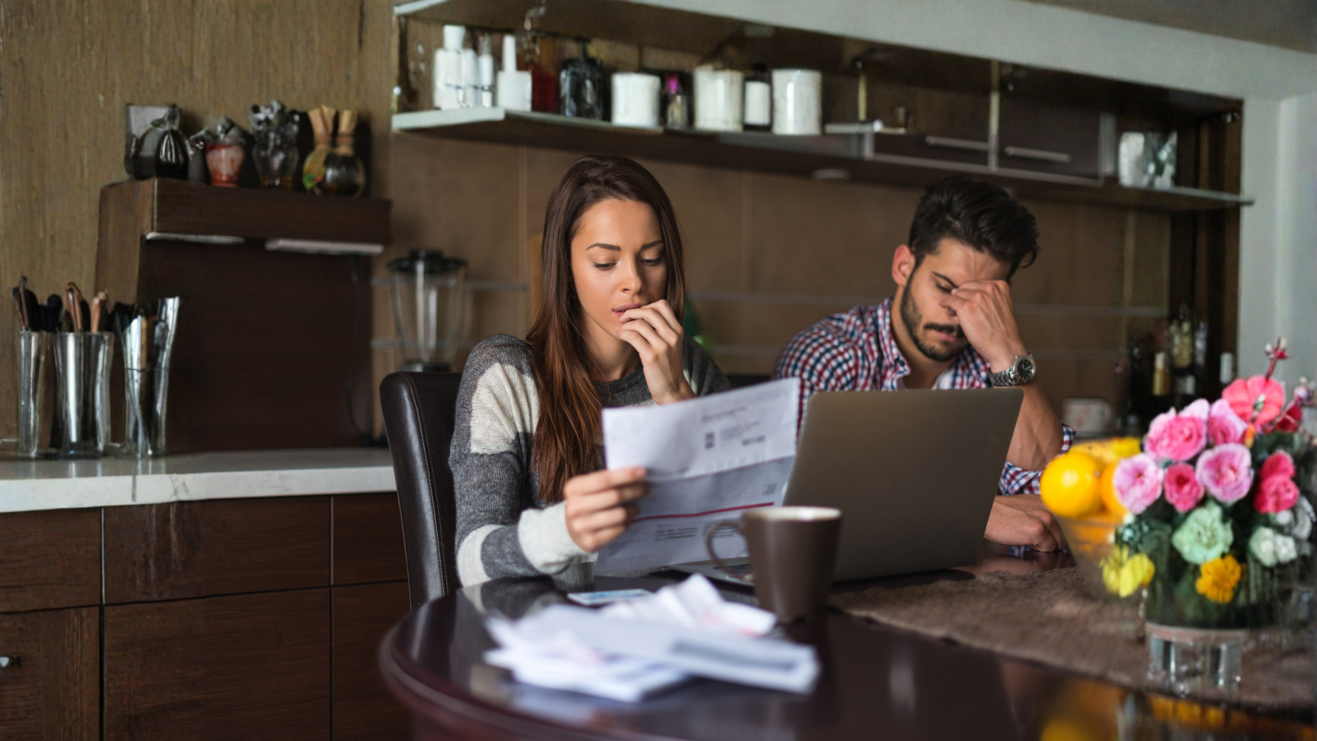 Two people sitting at a table, reading papers in a kitchen with coffee, fruit, and flowers.