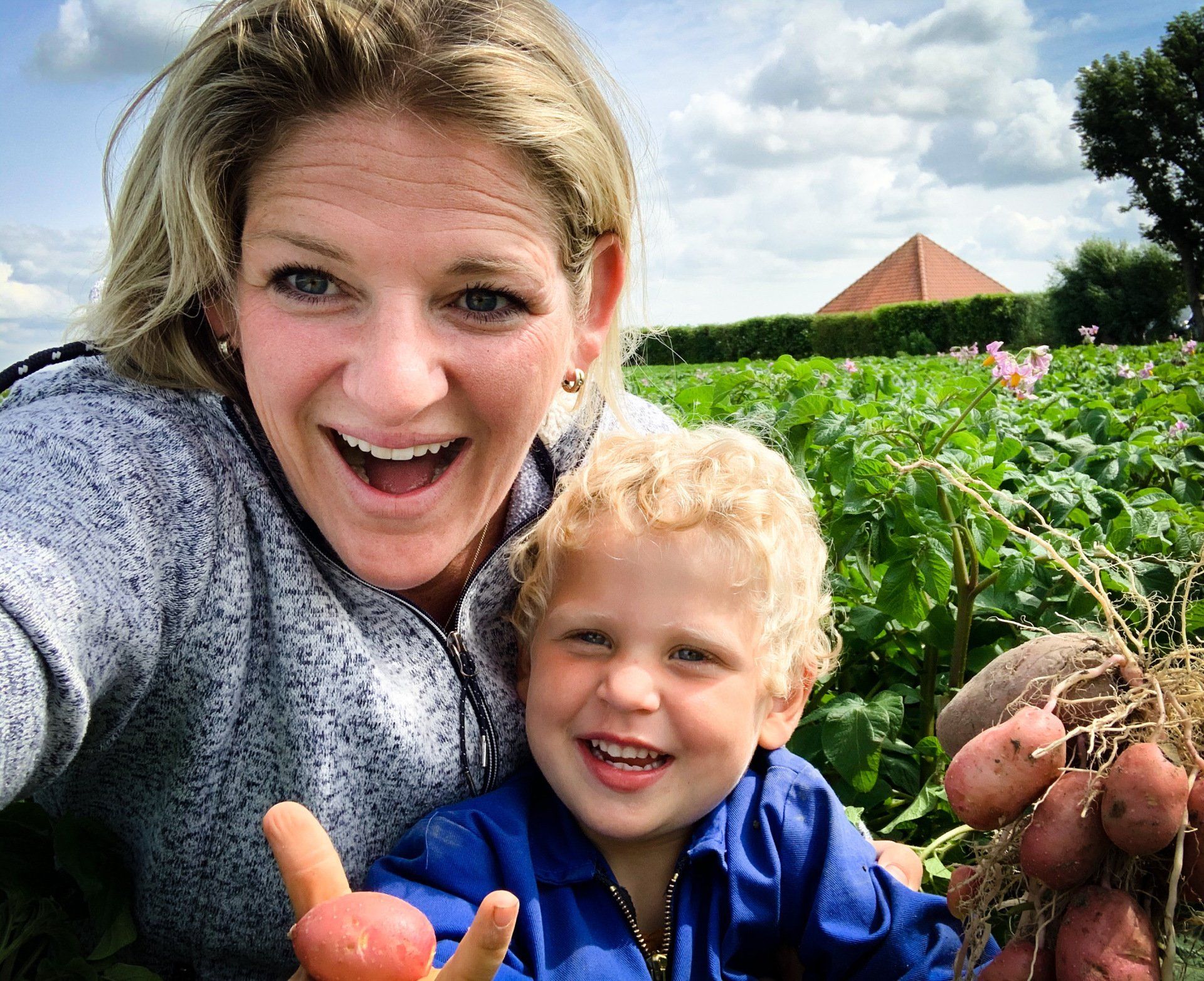 Vrouw en kind glimlachen in een veld, met vers geoogste aardappelen in hun handen.