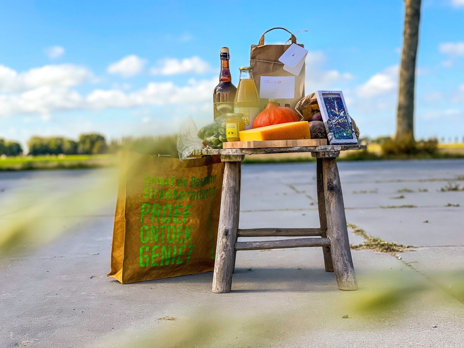 Een picknickmand, wijn, fruit en een klein bordje op een krukje langs een pad aan het water met een geweven tas.