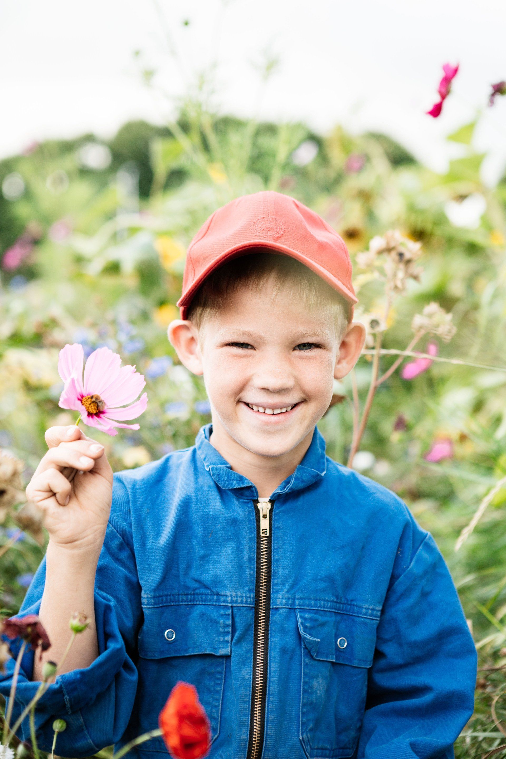 Een lachend kind met een rode muts en een blauwe jas houdt een roze bloem vast in een tuin.