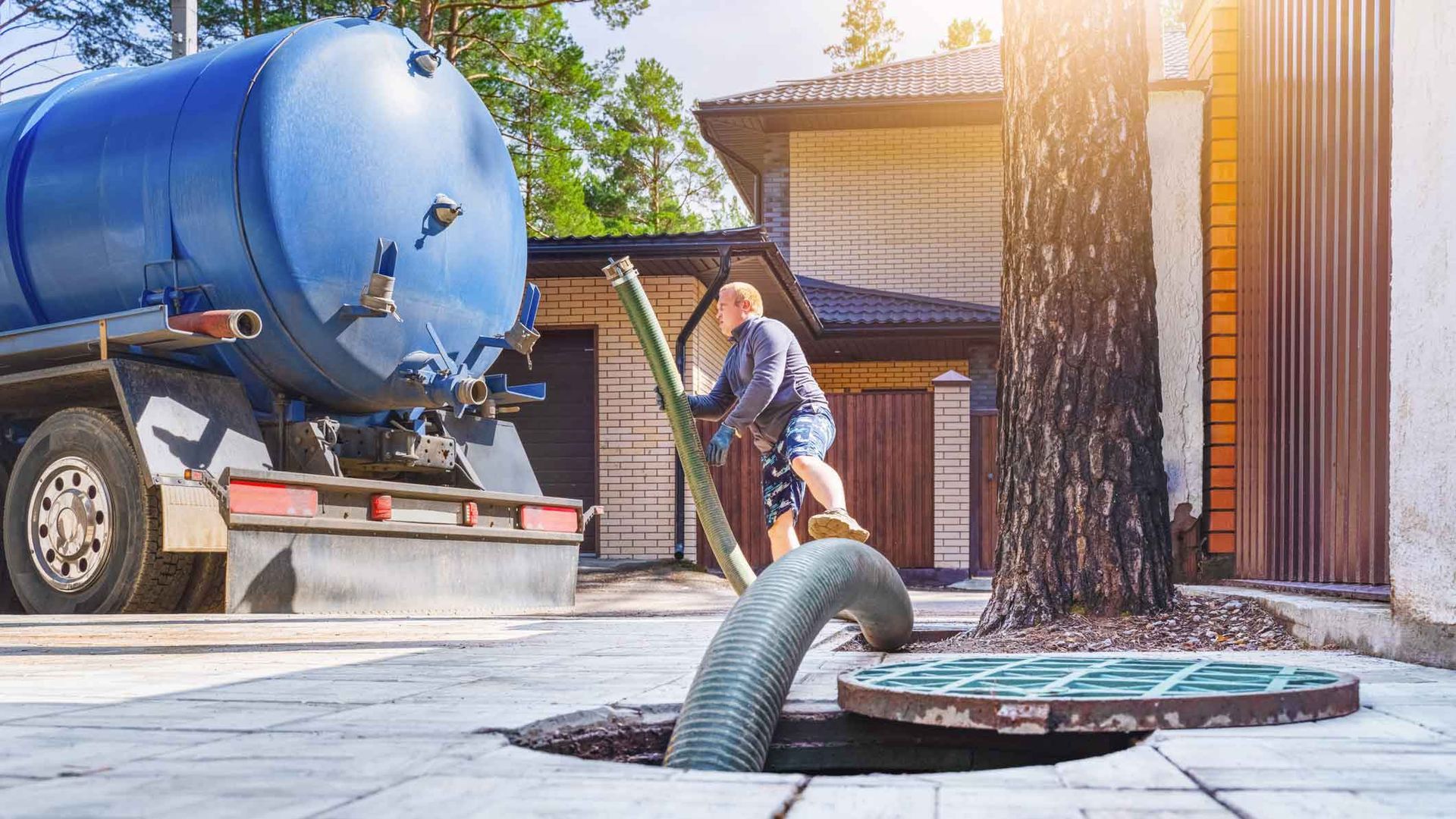 Technician guides a large suction hose into an open septic tank access lid on a concrete driveway.