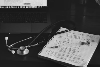 Stethoscope, notebook with handwritten notes, and laptop on a desk, in grayscale.