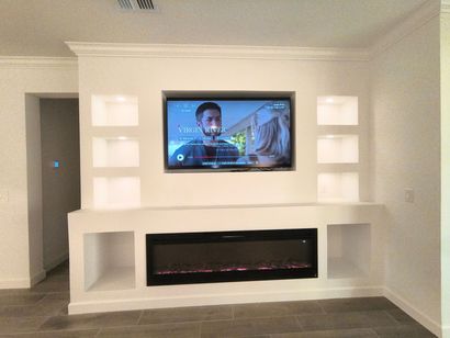 Built-in white wall unit with a mounted TV above an electric fireplace, flanked by lighted shelves.