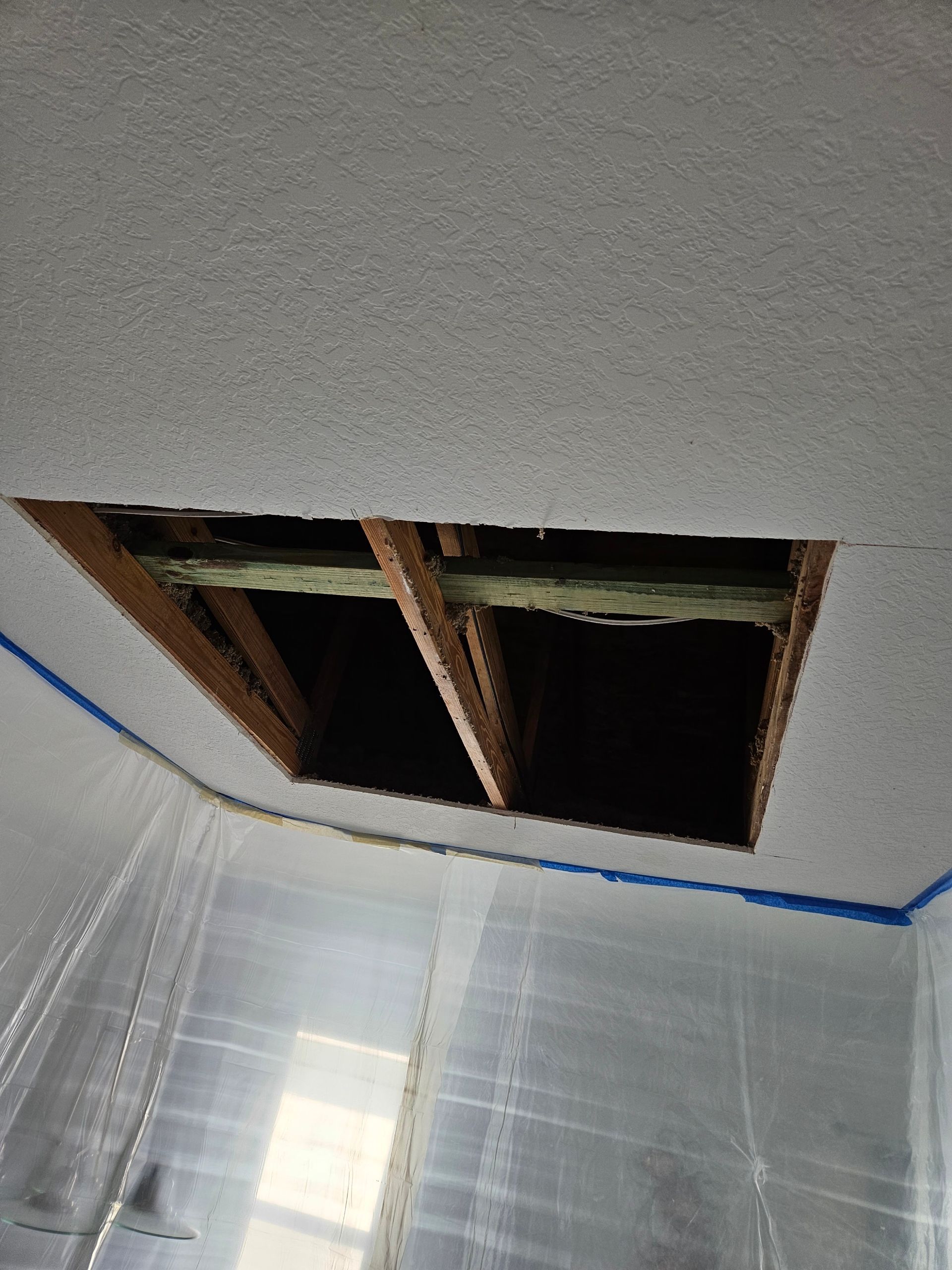 Hole in ceiling, revealing attic space. Wooden frame visible. Room is covered in plastic sheeting.