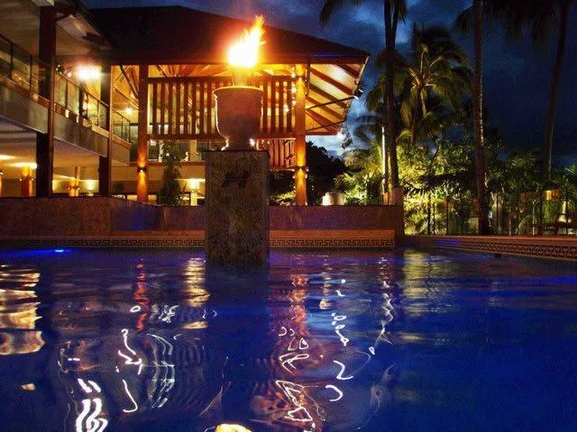 Nighttime resort pool with a lit thatched pavilion and reflections on the water