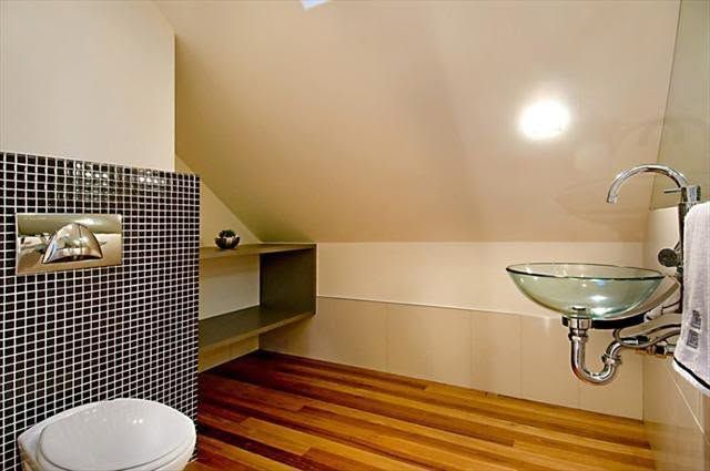 Small bathroom with wood floor, tiled wall, wall shelf, and glass sink under sloped ceiling.