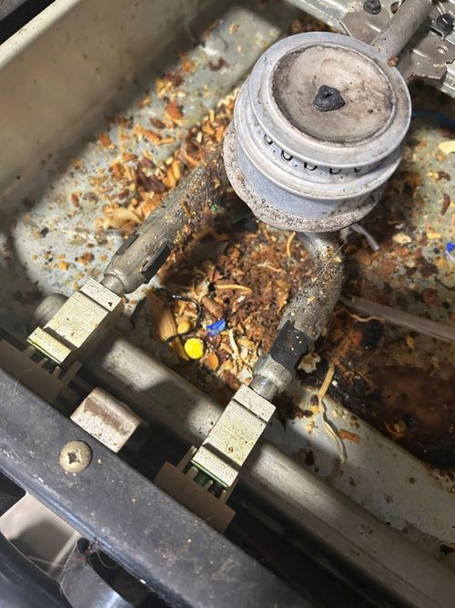 Close-up of a dirty, grease-stained stovetop burner with metallic parts and food debris accumulated underneath.