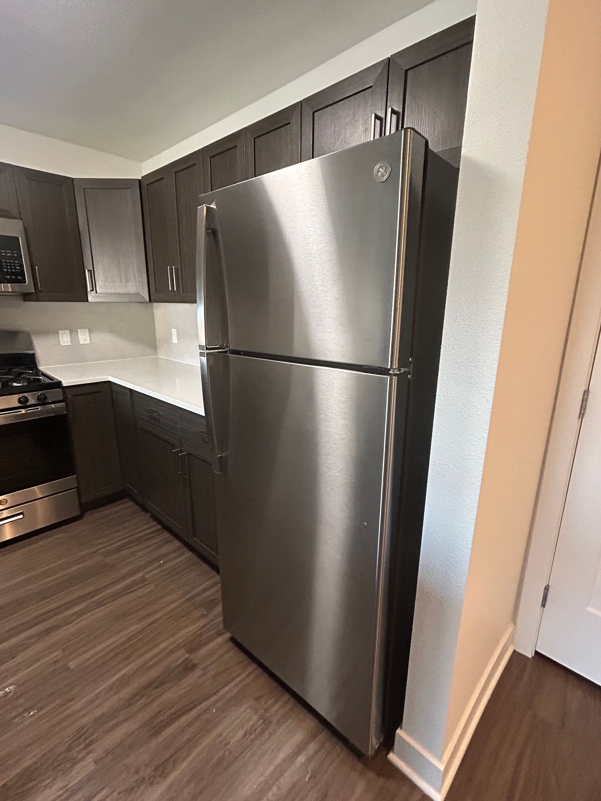A stainless steel top-freezer refrigerator in a modern kitchen with dark brown cabinets and wood-style flooring.