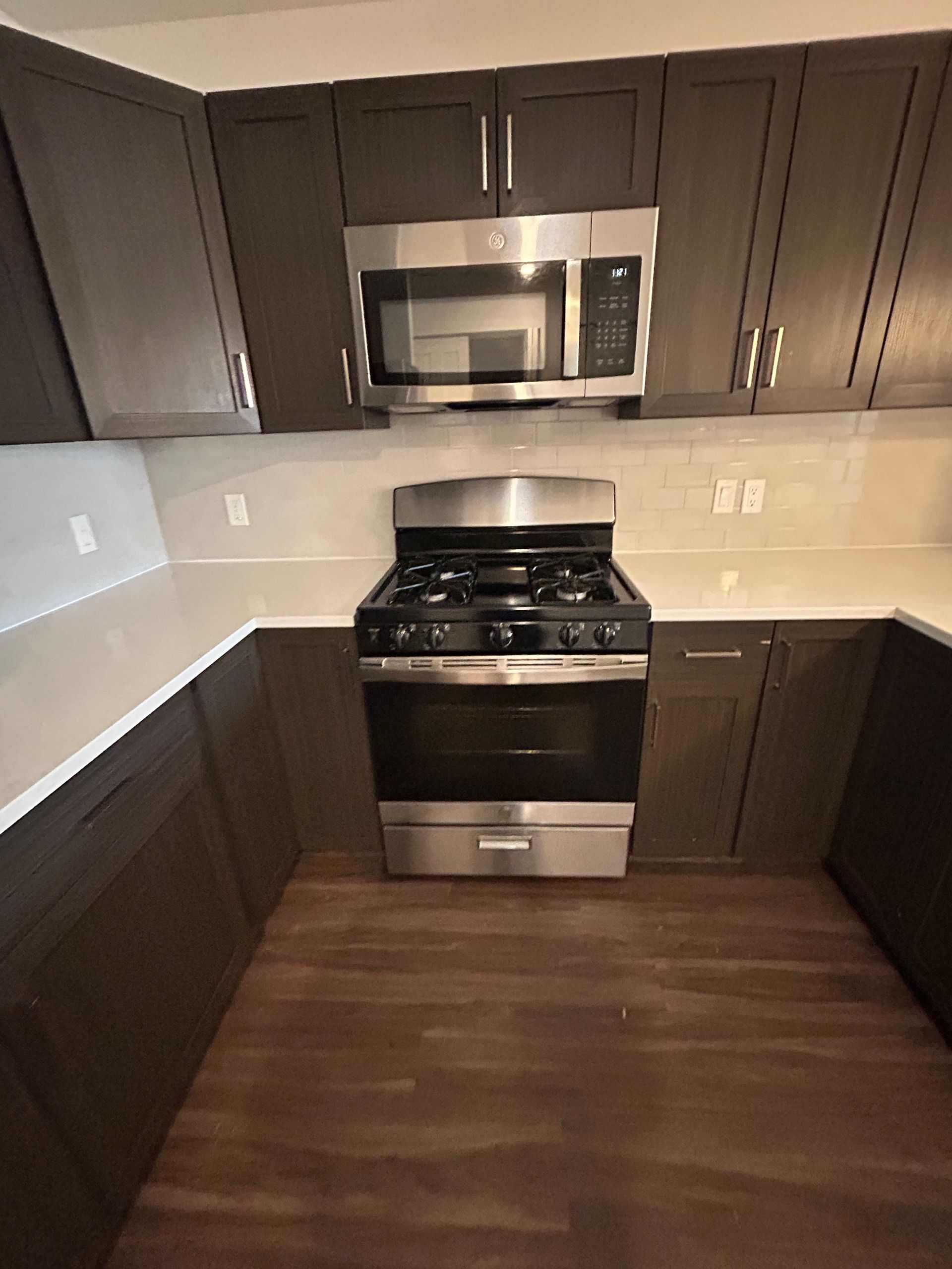 A modern kitchen with dark brown cabinets, white countertops, stainless steel stove, and an over-the-range microwave.