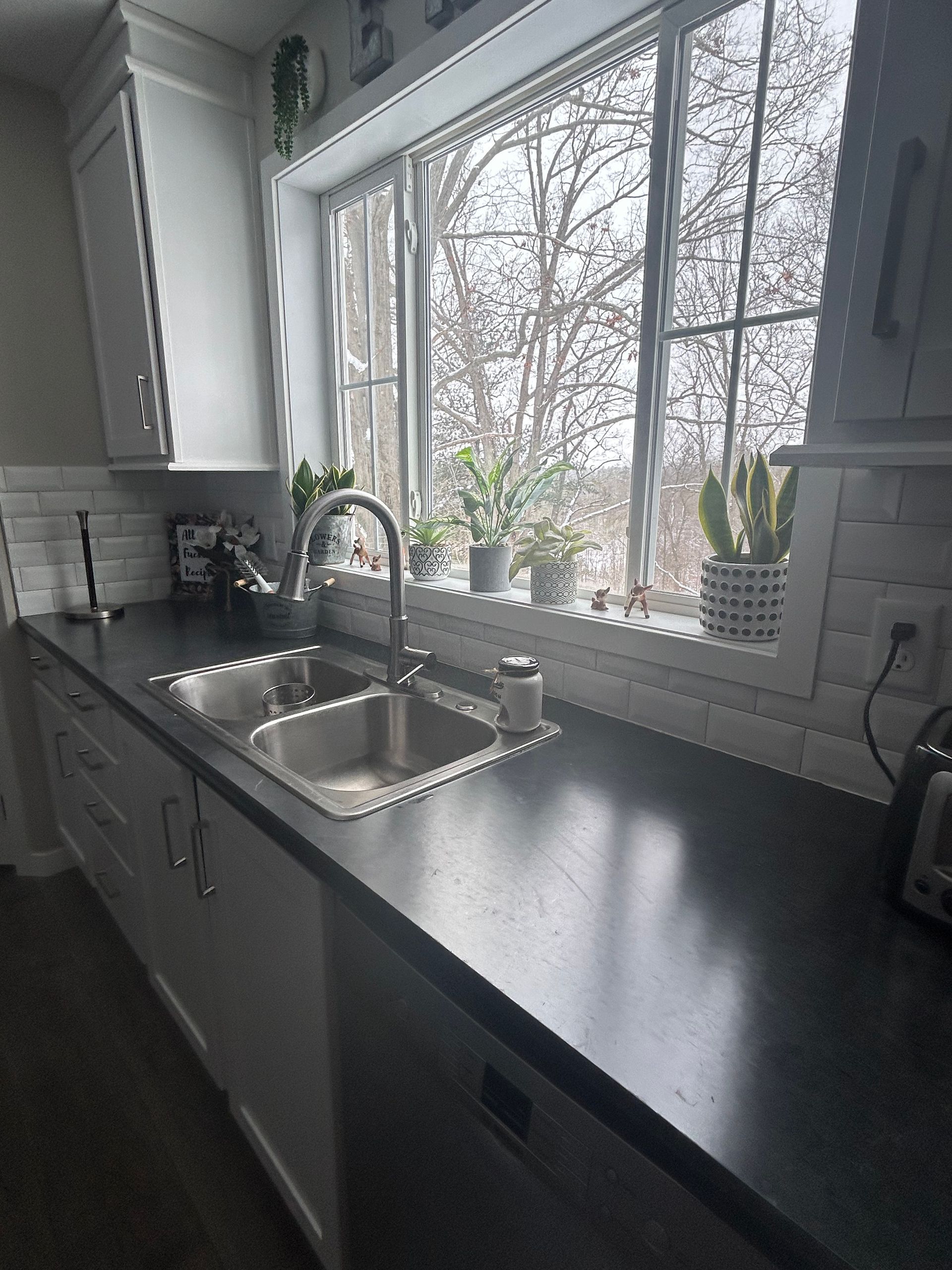 A kitchen sink with a chrome faucet set in a dark countertop under a large window filled with small potted plants.