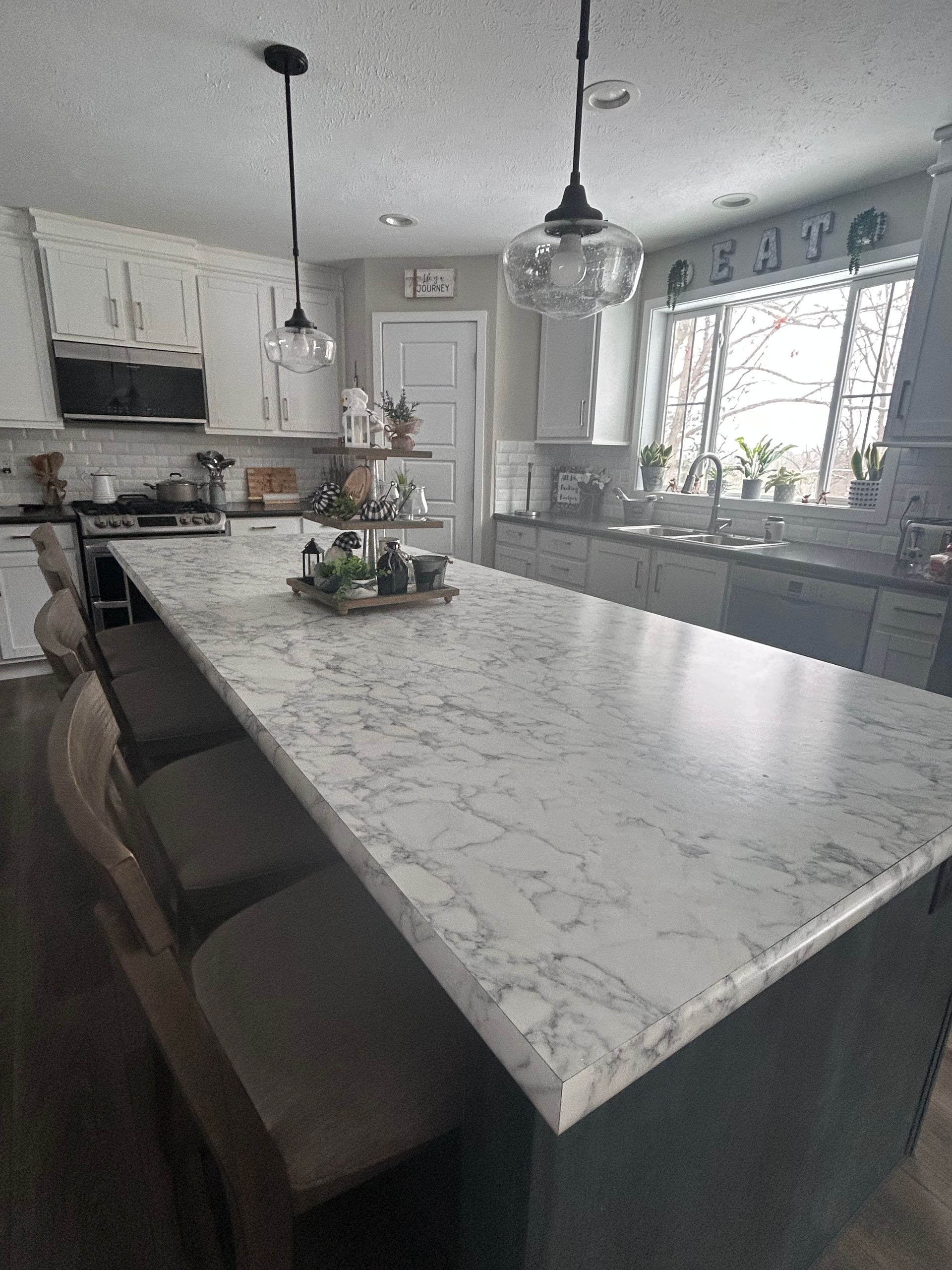 A kitchen island with a white marble countertop and stools, positioned in front of cabinets and a window.