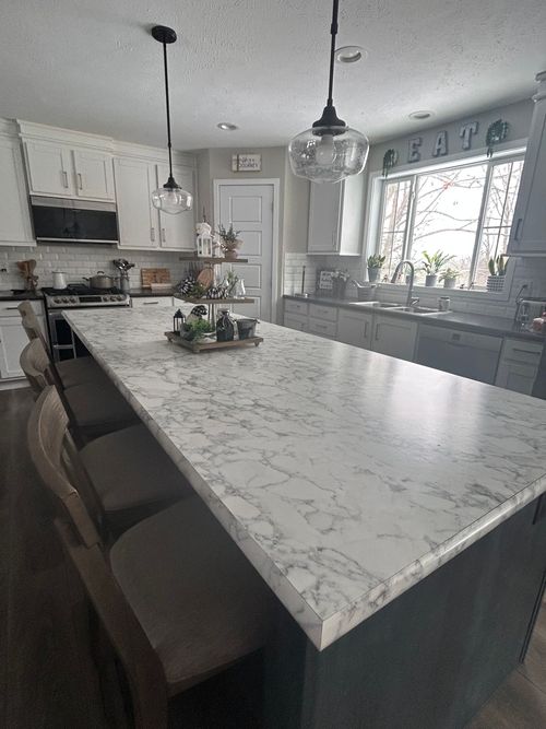 A kitchen island with a white marble countertop and stools, positioned in front of cabinets and a window.
