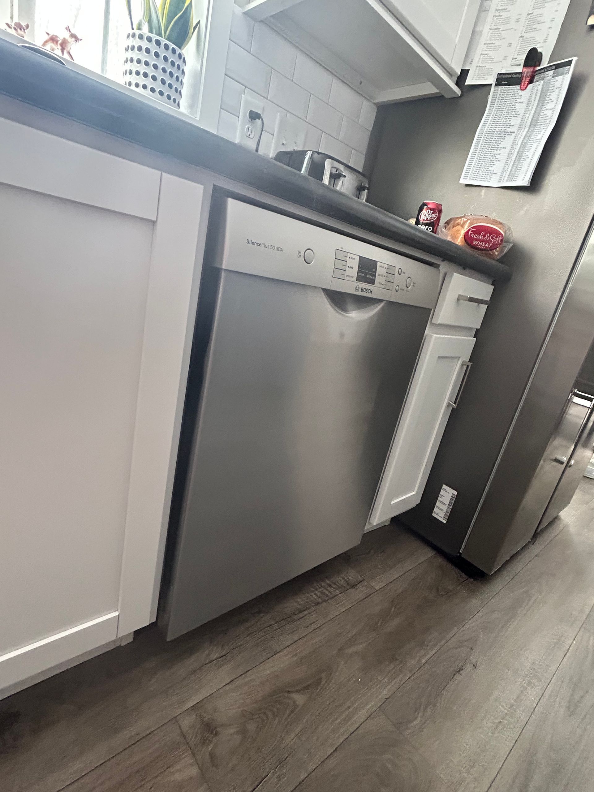 A stainless steel dishwasher is installed between white kitchen cabinets under a dark countertop in a home kitchen.