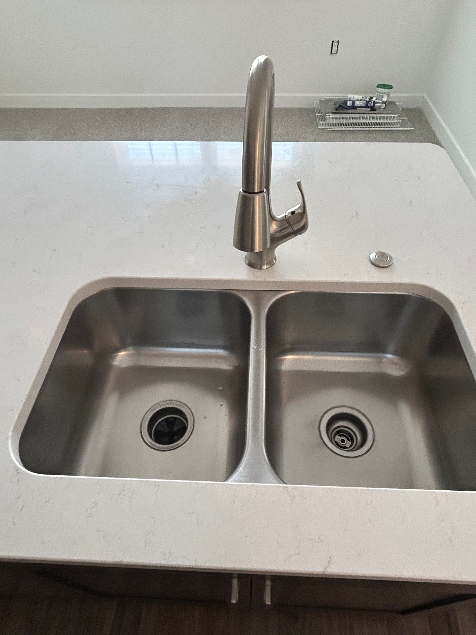 A double-basin stainless steel kitchen sink set into a white quartz countertop with a tall, modern brushed-nickel faucet.