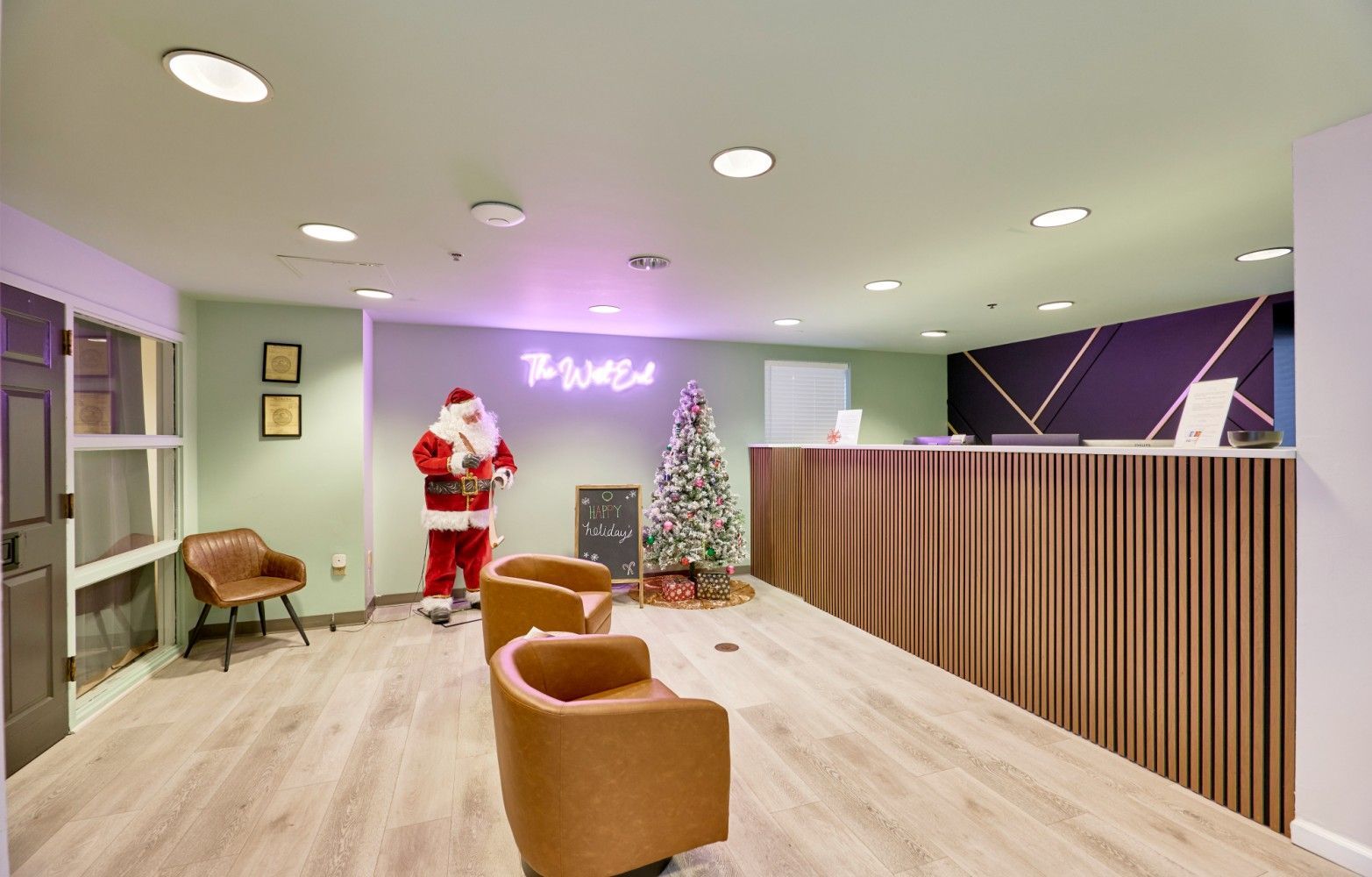 Bright lobby with Santa by a decorated tree, tan chairs, and a wooden reception desk under purple lighting