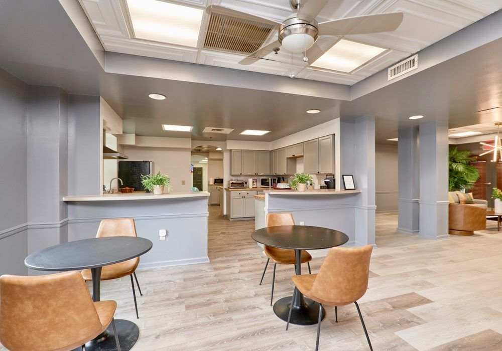 Bright lobby seating area with round tables, tan chairs, and a reception desk under a ceiling fan and skylights