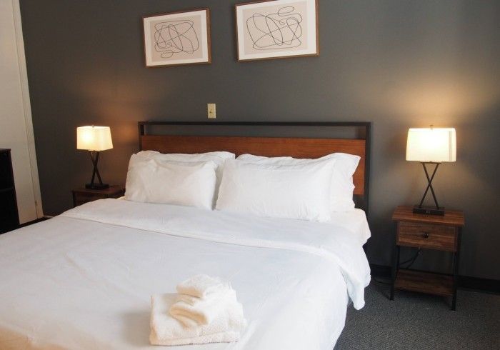 Neatly made bed in a dark-walled bedroom with two bedside lamps and framed art above the headboard