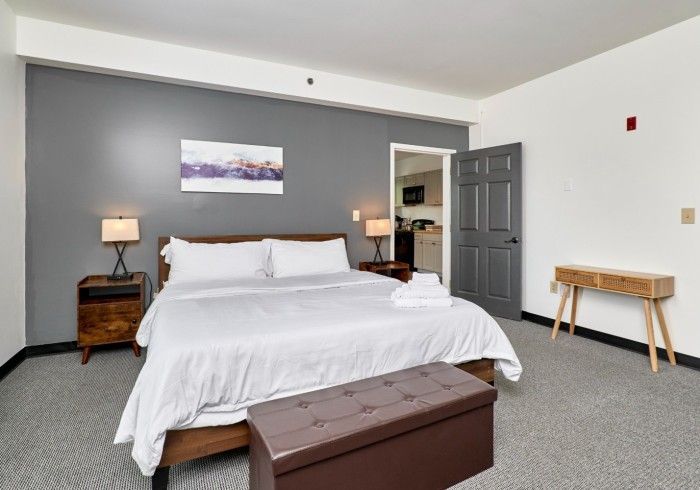 Spacious bedroom with a large bed, gray walls, bedside lamps, and a small wooden table near the door.