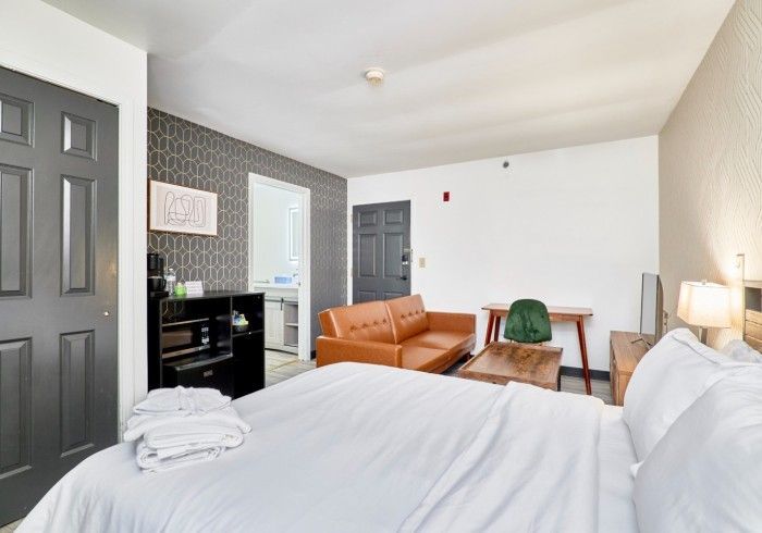 Bright hotel room with bed, orange sofa, small table, and dark gray door near a window.